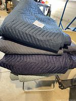 Four heavy duty furniture moving blankets stacked, showing various dark colors and zigzag quilted pattern, with wear visible.