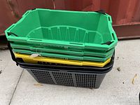 Stack of four green plastic grocery baskets with black handles on top of three black perforated baskets.