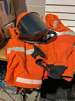 Orange electrical safety jacket with reflective stripes and attached helmet with clear face shield placed on a bench.