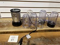 Magic Bullet blender base with multiple clear plastic mixing cups and blending blade, all placed on wooden surface against white slat wall background.