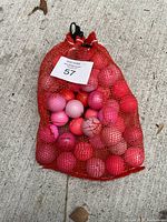 Red mesh bag filled with pink golf balls in various shades and brands, placed on concrete ground.