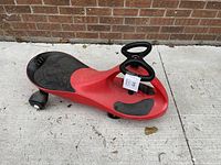 Red plasma car ride-on toy with visible wear placed on a concrete surface in front of a brick wall. Steering wheel in bowtie shape and wheels visible.
