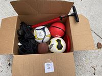 Viewed from above showing the box contents including two soccer balls, mini football, red mini basketball, black protective pads, red air pump, and red cone base.
