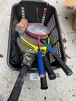 Top view of black basket containing four rackets and a container of orange tennis balls.