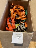 Box filled with multiple nylon tie straps in orange and other colors, showing metal ratchet buckles and hooks.