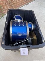 View of children's blue toy drum set inside a black storage bin, showing bass drum, two tom drums, and cymbal with foot pedal.