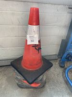 Single orange traffic cone with white reflective band on black square base and minor marks