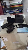 Black Nikon Action EX 7x35 waterproof binoculars on a cabinet with case, strap, and manual, books in background.