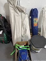 Two beige carrying bags, blue carrying bag, box with camping items and a bucket seen against wall; variety of camping gear seen.