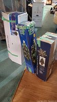 Three boxed artificial Christmas trees; one shows an image of a 32-inch fibre optic tree with snowflakes on the box.