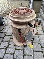 Front and side views of an outdoor gas fire pot with beige textured finish and diamond pattern. Top is filled with reddish-brown lava rocks surrounding central burner. Includes red and green straps with cords for carrying.