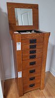 Tall wooden jewelry armoire with 8 drawers and black metal handles, mirror inside lid visible with some jewelry inside compartment.