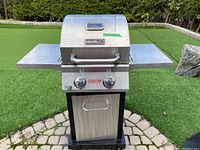 Full view of NexGrill Evolution propane BBQ grill with two side shelves set up outdoors on paving stones next to artificial grass.