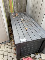 Side view of the rectangular Keter outdoor storage box with slatted top and panel sides, showing some outdoor dirt and leaves on top.