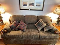 Full front view of three-seat sofa with brown textured upholstery, wood trim on arms, and four throw cushions (two floral, two plaid).