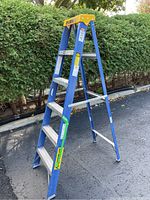 Side angle showing the 6 foot blue fiberglass frame step ladder with yellow top and aluminum steps on paved driveway.