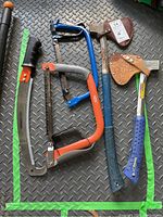 Photo of two axes and three saws laid out on a metal textured surface, showing handles, blade covers, and sheaths.