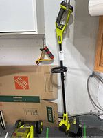 Full view of the Ryobi battery operated trimmer leaning against a wall with cardboard boxes behind it.