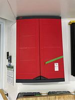 Closed front view of red Keter wall storage unit mounted on wall with two doors and black trim, showing green tape securing it.