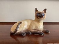 Side profile of the ceramic Siamese cat figurine lying down, showing cream body and dark brown limbs and tail.