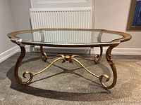 Front angle view of the gold gilt metal coffee table with an oval glass top showcasing the intricate scrolled legs and crossbar supports.