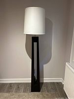 Front view of tall black floor lamp with open rectangular base and white drum shade against a wall on wood flooring.