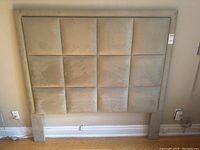 Front view of a queen size light beige ultrasuede headboard with 12 padded panels and two attached legs.