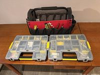Photo showing soft-sided black and red tool bag with reinforced handle and multiple exterior pockets containing hand tools. Two clear compartment organizers with yellow latches filled with screws and hardware in separate compartments visible in front of the bag.