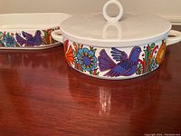 Two Villeroy & Boch Acapulco pattern serving dishes on a wooden table, one round with lid and one oval without lid, both with colorful floral and bird designs.