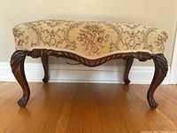 Side view of footstool showing carved cabriole wooden legs, wood frame with carved rope detail, and tapestry upholstered cushioned top