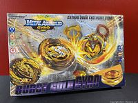 Front of sealed box showing gold metal assembled gyro Beyblade toy with battle effect imagery and branding.