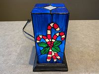 Front view of cube-shaped stained glass light with candy cane and green bow design on blue background, mounted on a black square base.