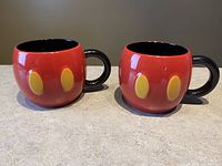 Photo showing front and side view of two Mickey Mouse collectible mugs highlighting the red exterior with yellow oval buttons and black handles.