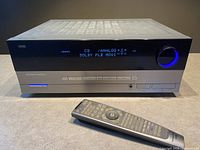 Front view of Harman Kardon AVR 144 receiver powered on with blue display, showing CD/ANALOG and Dolby PLII MOVIE, with remote control placed in front.