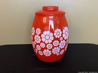 Front view of a red ceramic cookie jar with white daisy floral pattern on the body and a matching lid with a knob on top.