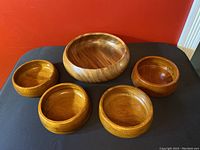 All five teak wood salad bowls arranged on a black cloth with orange background wall, showing natural wood grain and warm brown tones.