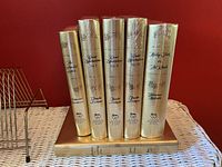 Front view showing six sealed gold-colored hardcover books standing upright and one laying flat, titles and authors fully visible on spines.
