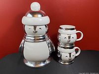 Set of one large penguin cookie jar and two smaller penguin mugs stacked together against a red background on black surface.