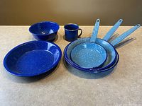 Alternate angled view of the 6-piece vintage blue enamelware set on a gray surface, presenting the handle details on the frying pans and visibility of all items.