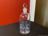 Full view of clear glass apothecary bottle with stopper and label Acid Acetic HC2H3O2 in white text.