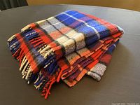 Folded vintage Ayers tartan wool blanket in red, blue, white, and beige colors with visible fringe tassels.