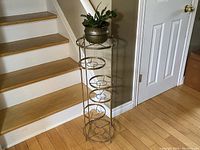 Full view of tall brass tiered plant stand with a plant placed on top tier, positioned next to stairs.