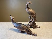 Two resin peacock figurines shown on a flat surface against a plain background, one in an upright elegant posture and the other in a lower position with detailed bronze-like finish.