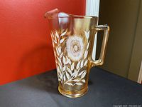 Amber iridescent carnival glass pitcher with white floral and leaf pattern, shown against a red background on black surface.