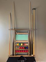 Full view of the pool cue rack with pool cues inserted and pool balls arranged on shelves, with scorekeeper board in the middle.