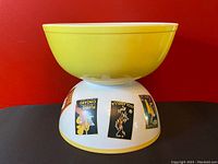 Photo of the bright yellow Pyrex Primary Yellow mixing bowl model 404 stacked on top of the white Galleria serving bowl with floral labels, showing the underside of the Galleria bowl with labels visible.
