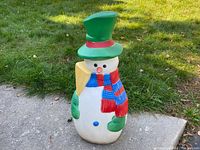 Front view of the blow mold vintage snowman showing the green top hat, carrot nose, red and blue scarf, green mittens, and blue button.