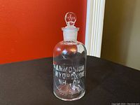 Full view of the clear vintage glass apothecary bottle with white embossed lettering 'Ammonium Hydroxide NH4OH'. Bottle is about 5 inches tall, standing on a dark surface against a red and beige background.