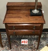Front view showing two-drawer writing table with hinged top compartments and brass drop ring pulls on a patterned rug with lamp placed on top.