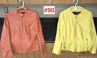 Two leather jackets, one light brown and one yellow, hung on white hangers against a black countertop background.